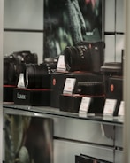 A display case with various camera equipment, including cameras and lenses, is arranged on shelves. Price tags are placed in front of the items, indicating an electronics or camera store setting. The equipment is prominently branded with a particular logo.