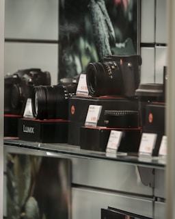 A display case with various camera equipment, including cameras and lenses, is arranged on shelves. Price tags are placed in front of the items, indicating an electronics or camera store setting. The equipment is prominently branded with a particular logo.