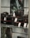 A display case with various camera equipment, including cameras and lenses, is arranged on shelves. Price tags are placed in front of the items, indicating an electronics or camera store setting. The equipment is prominently branded with a particular logo.