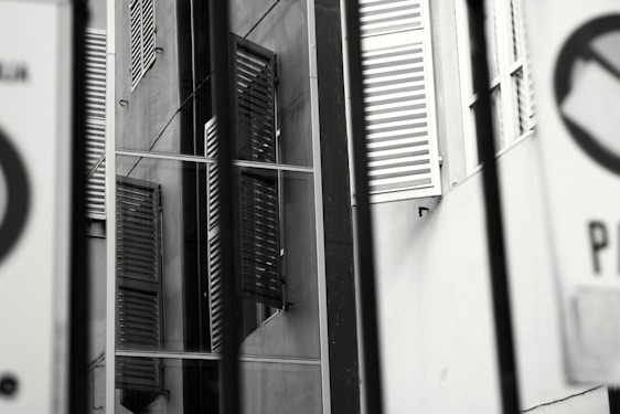 Multiple angled views of a building facade feature reflective windows and partially open shutters. Signs with symbolic graphics are partially visible, set against an urban backdrop of concrete and glass.