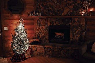 A cozy living room decorated for the holidays features a lit Christmas tree adorned with white lights and ornaments. The room has wooden walls and a rustic stone fireplace. Small candles are placed on the mantel along with decorative items like a small antler. A present wrapped in checkered fabric sits at the base of the tree.