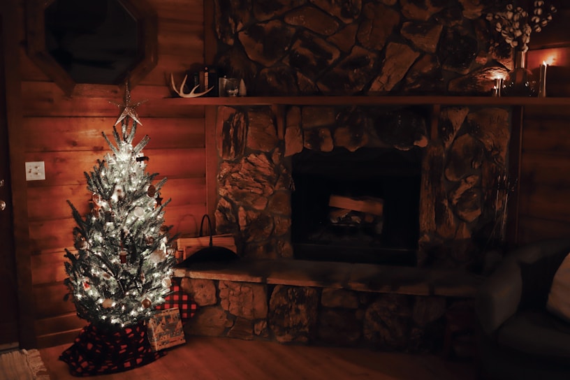 A cozy living room decorated for the holidays features a lit Christmas tree adorned with white lights and ornaments. The room has wooden walls and a rustic stone fireplace. Small candles are placed on the mantel along with decorative items like a small antler. A present wrapped in checkered fabric sits at the base of the tree.