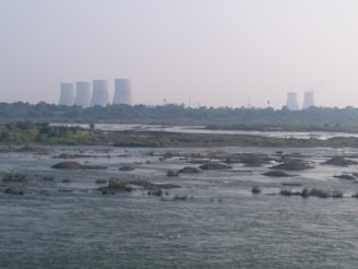 Cooling towers from a power plant are visible in the distance, with a river flowing in the foreground. The landscape is dotted with patches of greenery and small islands within the water. The sky is slightly overcast, adding a hazy appearance to the scene.