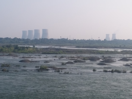 Cooling towers from a power plant are visible in the distance, with a river flowing in the foreground. The landscape is dotted with patches of greenery and small islands within the water. The sky is slightly overcast, adding a hazy appearance to the scene.