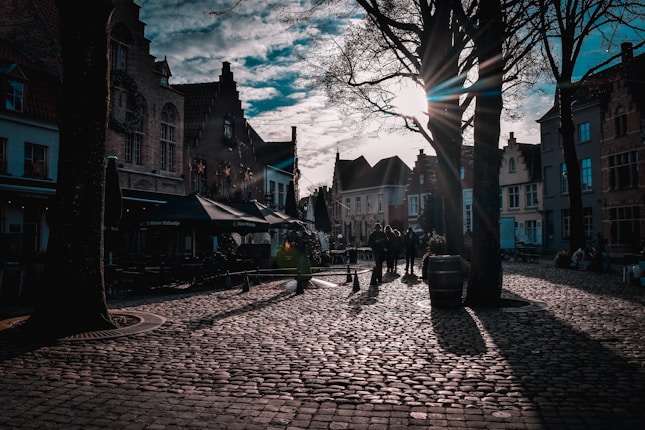 Cobblestone streets with historic European buildings create an old-world charm. Tall trees cast elongated shadows as the sun sets, radiating beams of light through the branches. People are casually walking and enjoying the picturesque environment.