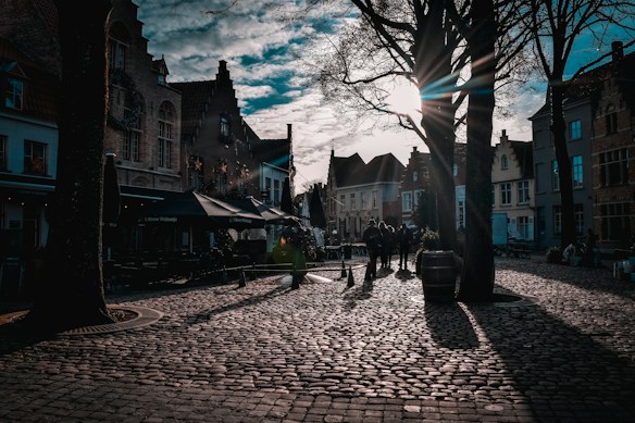 Cobblestone streets with historic European buildings create an old-world charm. Tall trees cast elongated shadows as the sun sets, radiating beams of light through the branches. People are casually walking and enjoying the picturesque environment.