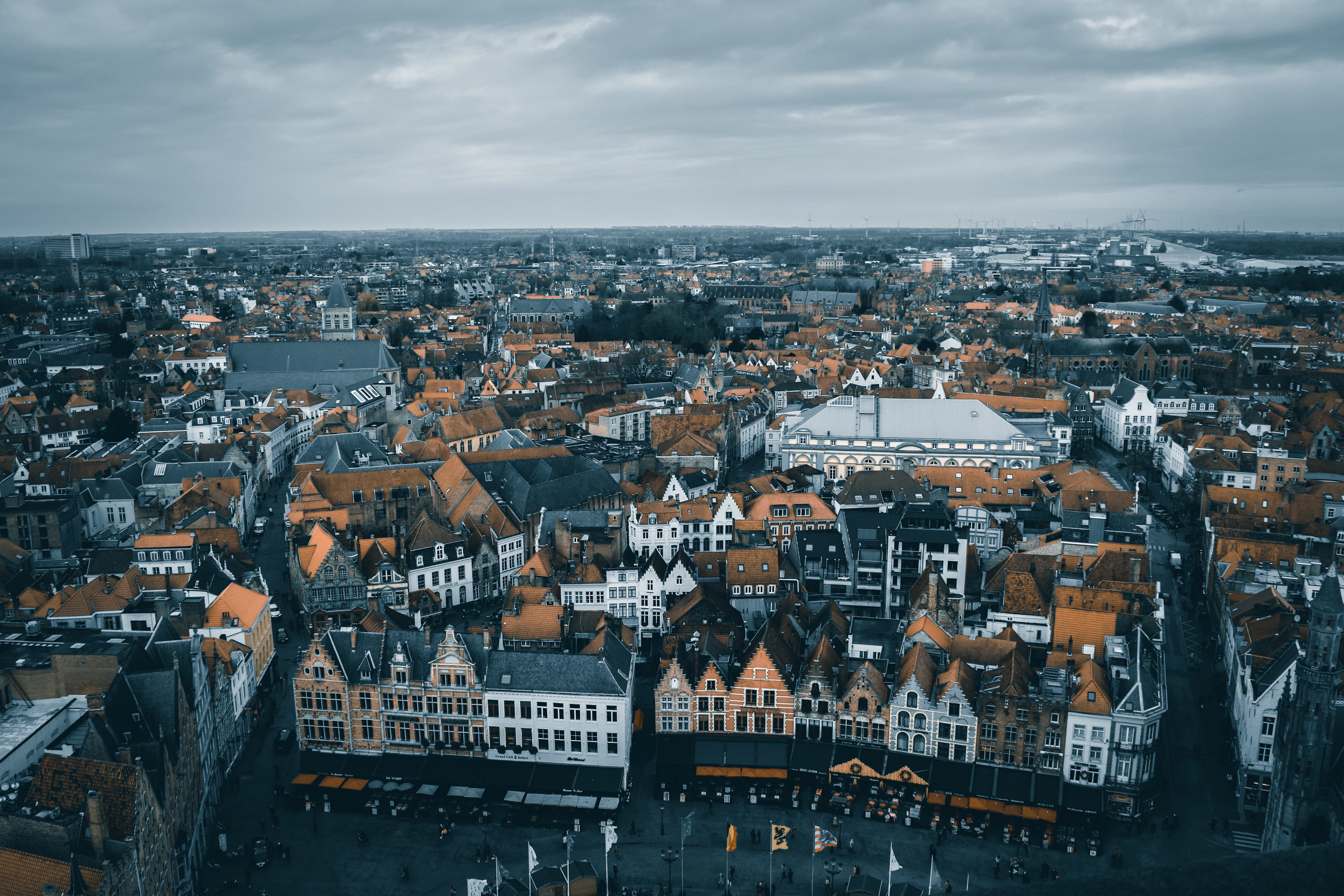 A moody aerial panorama over Bruges' red-tiled rooftops and market hall tower under a dramatic grey sky.