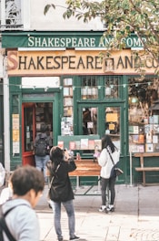 A quaint bookstore with a vintage exterior, featuring a prominent sign that reads 'Shakespeare And Company'. People are seen outside, some taking photos and others browsing books displayed on a table. The building has a green and gold color scheme with large windows exhibiting more books and posters.