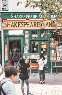 A quaint bookstore with a vintage exterior, featuring a prominent sign that reads 'Shakespeare And Company'. People are seen outside, some taking photos and others browsing books displayed on a table. The building has a green and gold color scheme with large windows exhibiting more books and posters.