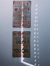Two motivational wall plaques are mounted on a white textured wall, illuminated by soft natural light. The top plaque features the words 'Dance, Love, Sing, Live' in bold red and black letters with smaller inspirational text beneath each word, along with music notes and a small heart icon. The bottom plaque features the text 'If you can imagine it, you can create it, if you dream it, you can become it' in red and black with varying sizes of font. The light creates a pattern of vertical, dotted shadows across the lower plaque.