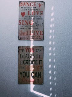 Two motivational wall plaques are mounted on a white textured wall, illuminated by soft natural light. The top plaque features the words 'Dance, Love, Sing, Live' in bold red and black letters with smaller inspirational text beneath each word, along with music notes and a small heart icon. The bottom plaque features the text 'If you can imagine it, you can create it, if you dream it, you can become it' in red and black with varying sizes of font. The light creates a pattern of vertical, dotted shadows across the lower plaque.
