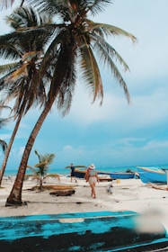 A tropical beach scene featuring tall palm trees swaying over a sandy shore. Two people are walking near colorful fishing boats on the sand, with one person in beachwear and a sunhat. The ocean in the background has vibrant blue hues, and the sky is partially cloudy.
