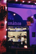 A cozy restaurant interior is viewed through a window adorned with decorative lights. Two people are in a kitchen area, engaged in culinary activities. The lighting casts a warm and inviting ambiance, with various kitchen utensils and equipment visible in the background. The restaurant is named 'Lunático'.