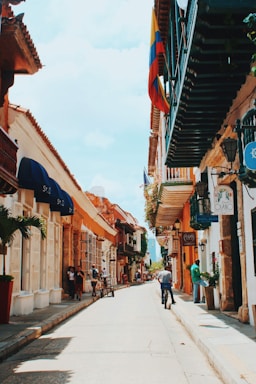 A narrow, vibrant street scene features colonial-style buildings with colorful facades and balconies adorned with flowers. A Colombian flag is prominently displayed, adding cultural significance. Pedestrians are casually walking or standing, while one person rides a bicycle down the sunlit street.