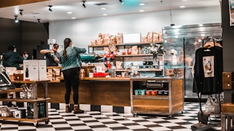 A cozy coffee shop with a black and white checkered floor hosts several people interacting at the counter. The counter is lined with various coffee products and merchandise, including bags of coffee and mugs. Shelves behind the counter are stocked with more items, and a refrigerator is visible on the right. A person in a green jacket and boots stands at the counter, while baristas prepare drinks.