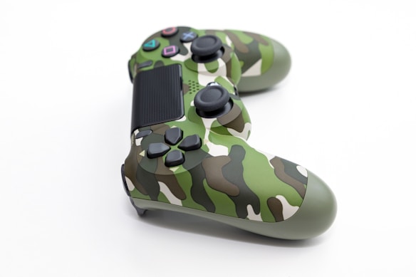 A game controller featuring a camouflage design with a mix of green, brown, and beige tones, set against a plain white background. The controller has multiple buttons and analog sticks, traditionally used for gaming consoles.