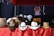 A collection of cowboy hats is arranged on a table covered with a red cloth that has musical notes printed on it. The hats vary in color and style, featuring white, brown, and black, some with decorative bands. In the background, there is a dark fabric backdrop with a white shirt hanging.
