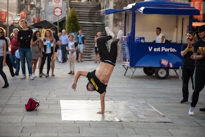 A person is performing a breakdance move, balancing on one hand with their legs in the air. A group of onlookers stand around watching the performance. The setting is an urban outdoor space with a blue vendor cart visible in the background.