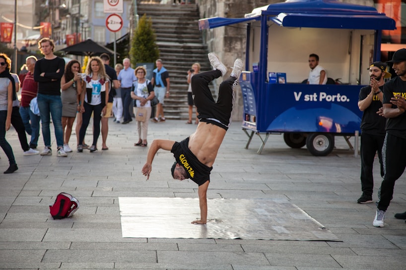 A person is performing a breakdance move, balancing on one hand with their legs in the air. A group of onlookers stand around watching the performance. The setting is an urban outdoor space with a blue vendor cart visible in the background.