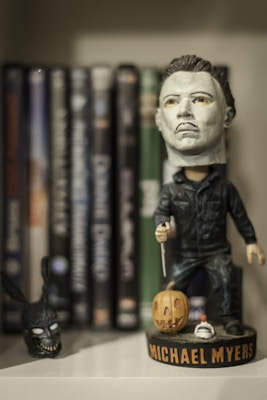 A figurine of a character stands in front of a shelf with DVDs. The figurine is holding a knife and dressed in a dark outfit, with a pumpkin at its feet. The name Michael Myers is on the base. A dark rabbit mask is placed to the left.