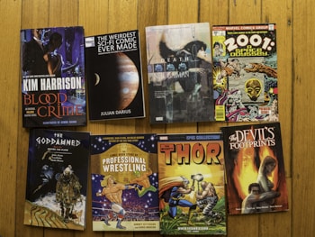 A collection of eight books and comic books are arranged on a wooden floor. The titles include a variety of genres such as fantasy, science fiction, crime, and nonfiction. Bright and colorful covers are displayed, featuring imagery ranging from planets and cosmic scenes to wrestlers and superheroes.