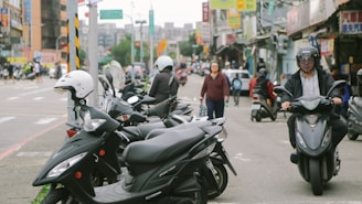 A busy urban street scene with multiple parked scooters lining the sidewalk. Several people are using scooters, and one person is riding towards the camera. There are various storefronts and signs in the background, indicating a bustling environment. A pedestrian is walking across the scene, and cars can be seen in the distance.