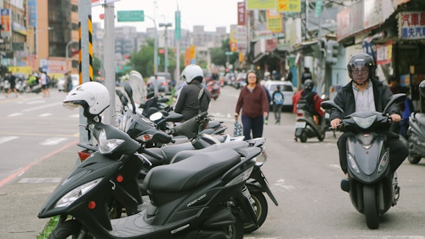 A busy urban street scene with multiple parked scooters lining the sidewalk. Several people are using scooters, and one person is riding towards the camera. There are various storefronts and signs in the background, indicating a bustling environment. A pedestrian is walking across the scene, and cars can be seen in the distance.