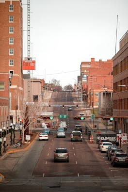 A view of an urban street with vehicles driving and parked along the sides. Tall brick buildings line the street, and traffic signals are visible above the road. A crane is attached to one of the buildings, possibly indicating construction work. The street sign indicates '5 ST', and the scene has a calm, regular city atmosphere.