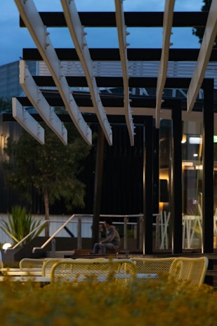 A modern outdoor seating area featuring geometric metal frames and tables is pictured. The area is surrounded by plants, and a person is sitting alone in the background against a backdrop of contemporary architecture. The scene is under dim lighting conditions, likely during dawn or dusk.
