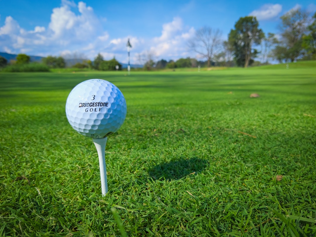 Golf ball on a tee with the hole flag in the background. Green grass and blue sky as the background. 