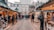A bustling outdoor market with wooden stalls lined up on either side of a wooden walkway. People are wearing winter clothing as they browse various goods, such as ornaments and crafts. Buildings and a monument are visible in the background, suggesting an urban setting. String lights and garlands add a festive atmosphere.