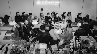 A black and white photograph captures a social gathering at a casual venue. Around a long, recessed table, a group of people sit closely together, engaged in conversation. Above them, a neon sign glows with the phrase 'Good to see you.' Hanging lamps illuminate the scene, creating a cozy atmosphere. In the foreground, a counter is visible with various cups and bottles, indicating a caf&eacute; setting.