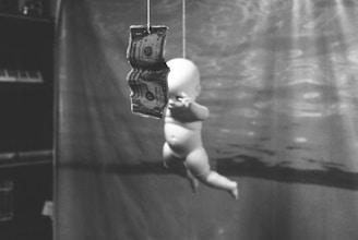A black and white photograph featuring a floating baby doll reaching towards a hanging dollar bill. The scene appears to be underwater, with light patterns suggesting sunlight filtering through water. The background is blurred, emphasizing the doll and the money.