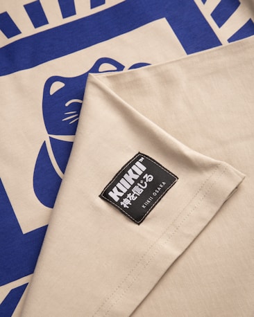 A fabric featuring a blue stylized cat design on a beige background, along with a sewn label that reads 'KIKI OSAKA' in both English and Japanese characters.
