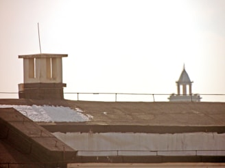 A rooftop with a chimney or vent structure partially covered in snow is in the foreground. In the distant background, a tower with a pointed roof is visible against a bright sky.