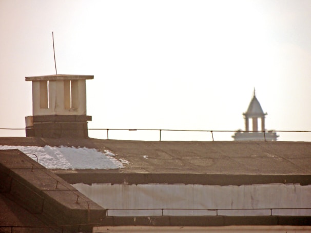 A rooftop with a chimney or vent structure partially covered in snow is in the foreground. In the distant background, a tower with a pointed roof is visible against a bright sky.