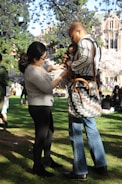 A couple is standing on a lush green lawn under blooming cherry blossom trees. The man is holding a baby, and the woman appears to be interacting with them. Other people are scattered in the background, enjoying the sunny day.