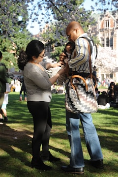 A couple is standing on a lush green lawn under blooming cherry blossom trees. The man is holding a baby, and the woman appears to be interacting with them. Other people are scattered in the background, enjoying the sunny day.