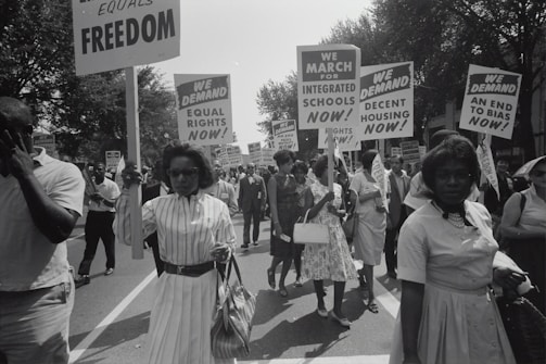 A group of people is marching down a street holding signs advocating for equal rights, integrated schools, decent housing, and an end to bias. The crowd appears to be diverse, and the march is peaceful with individuals dressed in mid-20th-century attire.