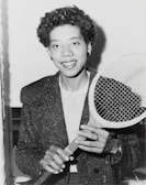A person holding a tennis racket appears in a black and white photograph. The individual is wearing a textured jacket over a white shirt and smiling at the camera. Their hair is curly, and they are accessorized with earrings and a ring.