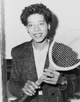 A person holding a tennis racket appears in a black and white photograph. The individual is wearing a textured jacket over a white shirt and smiling at the camera. Their hair is curly, and they are accessorized with earrings and a ring.