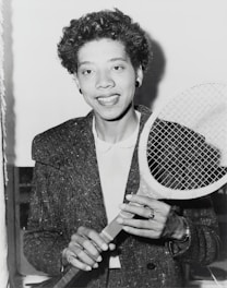 A person holding a tennis racket appears in a black and white photograph. The individual is wearing a textured jacket over a white shirt and smiling at the camera. Their hair is curly, and they are accessorized with earrings and a ring.