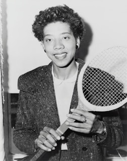 A person holding a tennis racket appears in a black and white photograph. The individual is wearing a textured jacket over a white shirt and smiling at the camera. Their hair is curly, and they are accessorized with earrings and a ring.