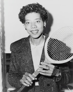 A person holding a tennis racket appears in a black and white photograph. The individual is wearing a textured jacket over a white shirt and smiling at the camera. Their hair is curly, and they are accessorized with earrings and a ring.