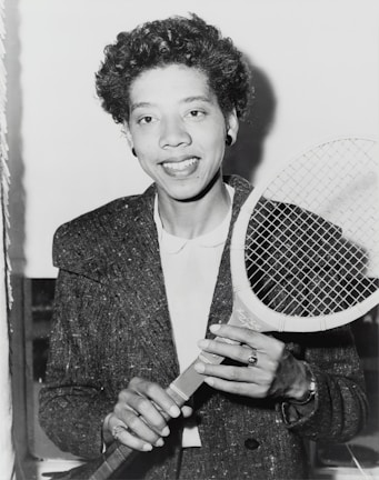 A person holding a tennis racket appears in a black and white photograph. The individual is wearing a textured jacket over a white shirt and smiling at the camera. Their hair is curly, and they are accessorized with earrings and a ring.