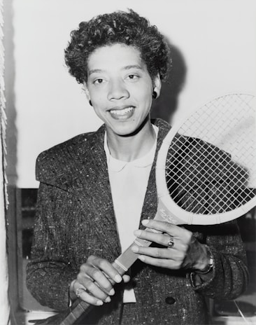 A person holding a tennis racket appears in a black and white photograph. The individual is wearing a textured jacket over a white shirt and smiling at the camera. Their hair is curly, and they are accessorized with earrings and a ring.