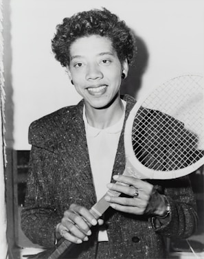 A person holding a tennis racket appears in a black and white photograph. The individual is wearing a textured jacket over a white shirt and smiling at the camera. Their hair is curly, and they are accessorized with earrings and a ring.