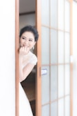 A bride stands partially behind a sliding wooden door with frosted glass panels. She is smiling warmly, with her hand touching her face, showcasing delicate earrings and a lace wedding dress. The setting appears serene and bright, with soft light illuminating her features.