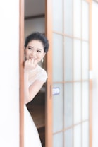 A bride stands partially behind a sliding wooden door with frosted glass panels. She is smiling warmly, with her hand touching her face, showcasing delicate earrings and a lace wedding dress. The setting appears serene and bright, with soft light illuminating her features.