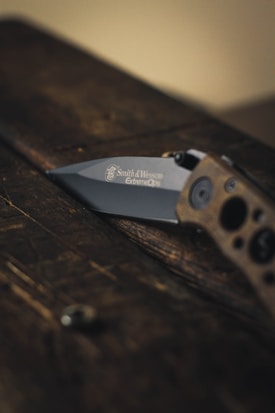 A folding knife partially open, resting on a dark wooden surface. The knife handle appears textured and the blade is engraved with the brand 'Smith & Wesson Extreme Ops'. The lighting is dim and atmospheric.